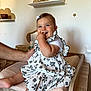 baby, child, smiling, sitting, dress, white, black_flower_pattern, hand, changing_table, room, clock, shelf, decor, star, fingers_in_mouth, happy, infant, cute, indoor, person