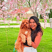 Tanuja Prabahar joined the competition — help win amazing prizes! woman, dog, pet, smiling, outdoor, spring, flower, cherry_blossom, green_grass, tree, nature, portrait, happy, curly_fur, holding, leash, urban, casual_clothing, daylight, backyard