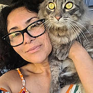 Bruce is registered to the contest to win money with this photo: woman, cat, selfie, glasses, yellow_eyes, curly_hair, tabby_cat, close_up, colorful_clothing, portrait, smile, indoor, face, pet, holding, shoulder, whiskers, feline, casual, friendly