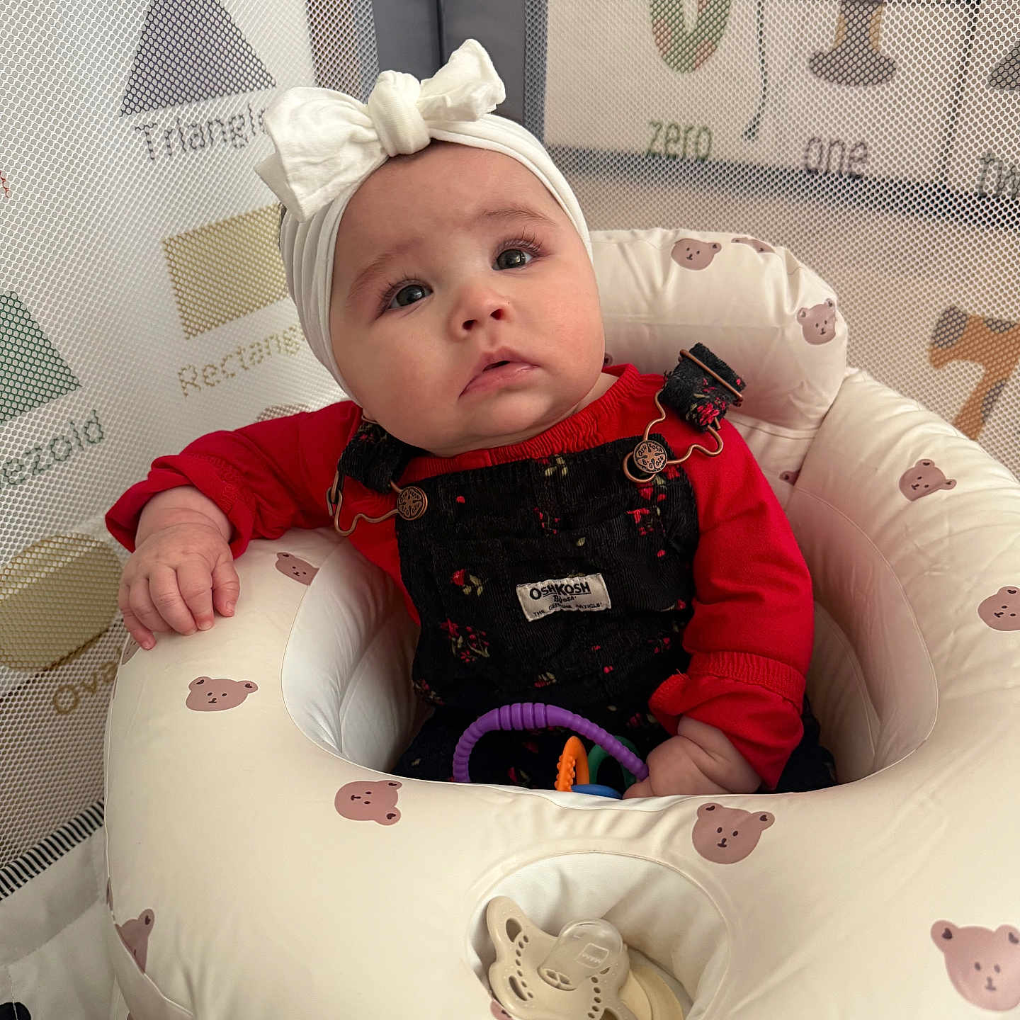 Nyla is registered to the contest to win money with this photo: baby, bear_pattern, bouncer_seat, bow, child, cute_expression, headband, indoor, infant, looking_up, mesh_net, number_blocks, overalls, pacifier, playpen, portrait, red_shirt, seat, shape_graphic, toy_rings