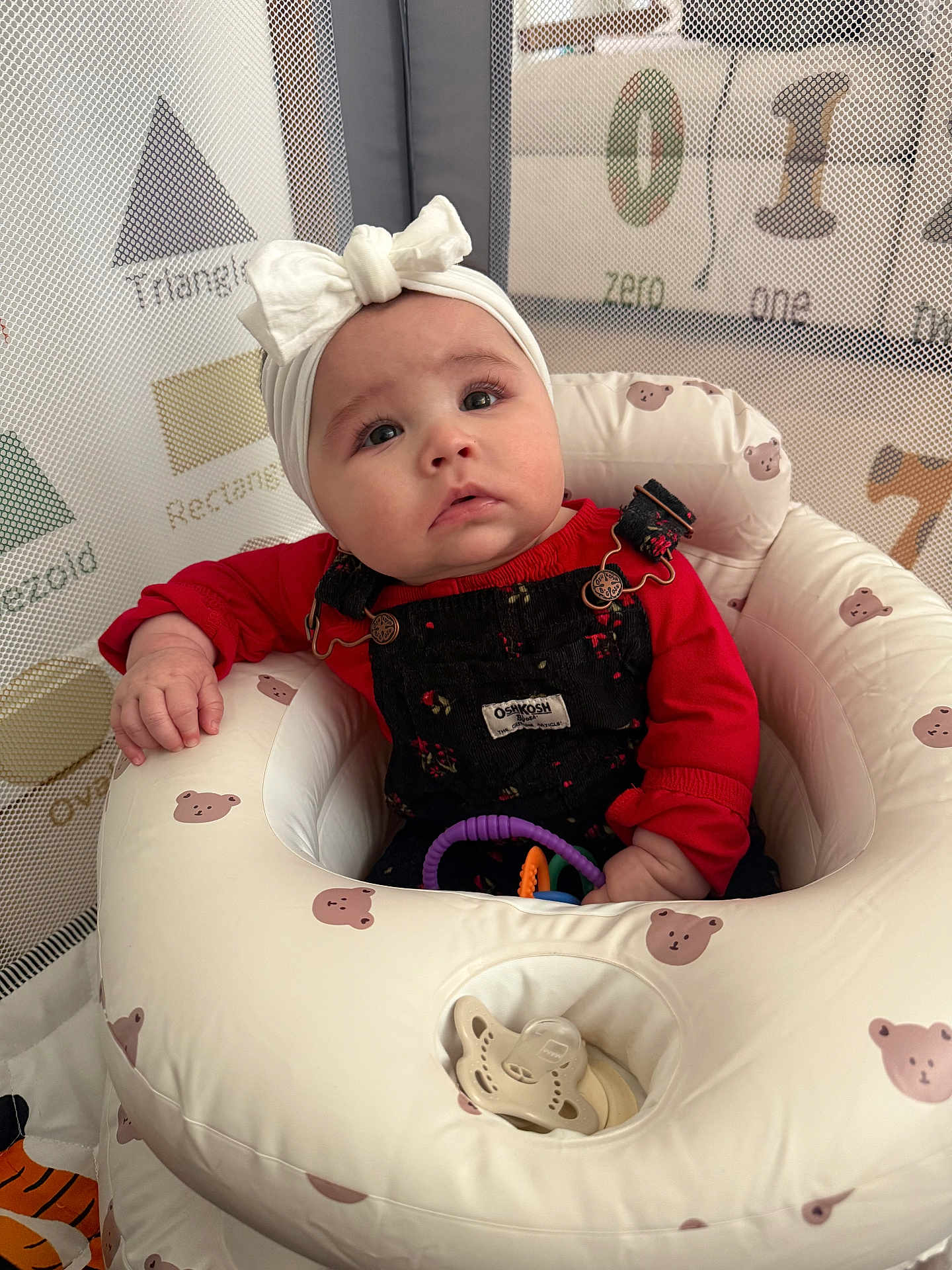 Nyla is registered to the contest to win money with this photo: baby, infant, headband, bow, overalls, red_shirt, bouncer_seat, toy_rings, pacifier, playpen, mesh_net, number_blocks, shape_graphic, bear_pattern, seat, portrait, looking_up, cute_expression, indoor, child