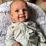 baby, infant, smile, face, eyes, onesie, clothing, hand, cushion, text, pattern, cute, indoors, portrait, child, happy, expression, soft, cozy, background