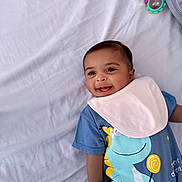 Nathaniel joined the competition — help win amazing prizes! baby, smiling, child, infant, blue_clothing, bib, dinosaur_print, toy, rattle, lying_down, white_sheet, happy, cute, portrait, young_child, indoors, soft_lighting, playful, closeup, face