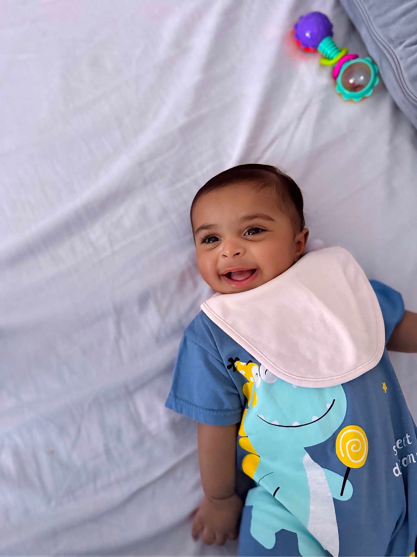 Nathaniel joined the competition — help win amazing prizes! baby, smiling, child, infant, blue_clothing, bib, dinosaur_print, toy, rattle, lying_down, white_sheet, happy, cute, portrait, young_child, indoors, soft_lighting, playful, closeup, face
