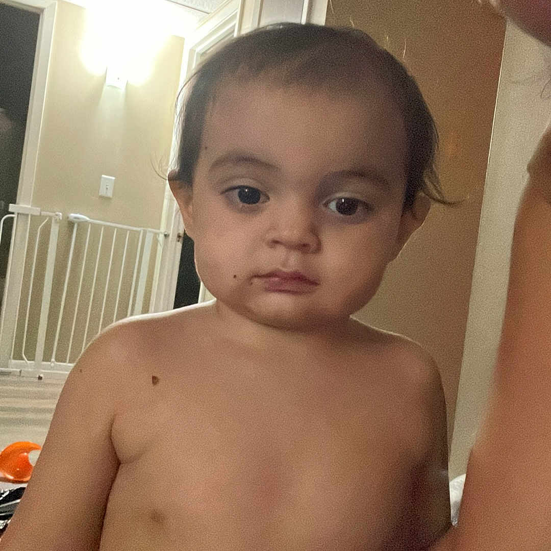 Kanye is registered to the contest to win money with this photo: toddler, child, baby, portrait, standing, indoors, home, hallway, bare_chested, face, eyes, hair, serious_expression, adult_arm, baby_gate, doorway, wall, ceiling_light, home_interior, photograph