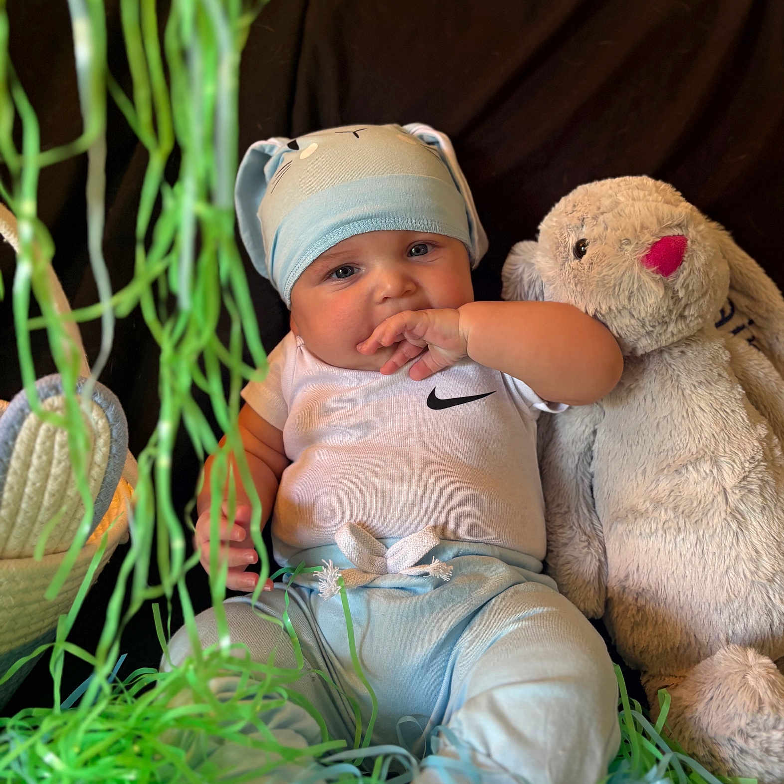 Kaine is registered to the contest to win money with this photo: baby, child, bunny_hat, stuffed_animal, plush_toy, blue_pants, white_shirt, green_shredded_paper, black_background, baby_sitting, hand_in_mouth, cute, indoor, soft_toy, hat, infant, portrait, playful, toy, colorful