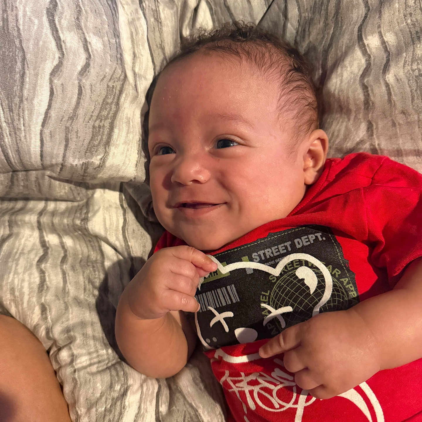 Zakyius is registered to the contest to win money with this photo: baby, smiling, red_shirt, bed, blanket, infant, face, hand, cute, lying_down, person, child, textile, bedroom, portrait, happy, skin, head, toddler, clothing