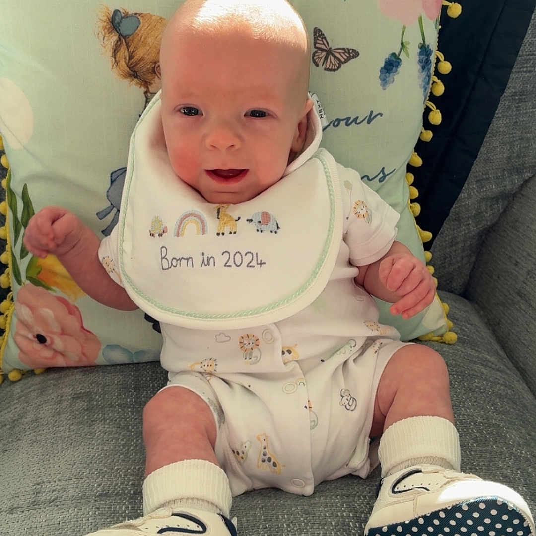 Franklin is registered to the contest to win money with this photo: baby, bib, child, clothing, couch, cute, face, footwear, happy, home, indoor, infant, newborn, patterned, pillow, shoes, sitting, smile, socks, soft_light