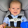 baby, child, infant, smiling, car_seat, seatbelt, harness, vehicle, car_interior, toy, remote_control, sippy_cup, blue_cup, blond_hair, portrait, indoors, travel, safe_seating, red_car, happy