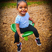 Aluna joined the competition — help win amazing prizes! child, girl, smiling, playground, green, hair_bow, curly_hair, sneakers, black_pants, blue_shirt, sitting, wood_chips, outdoor, daylight, happy, park, toy, play, person, cute