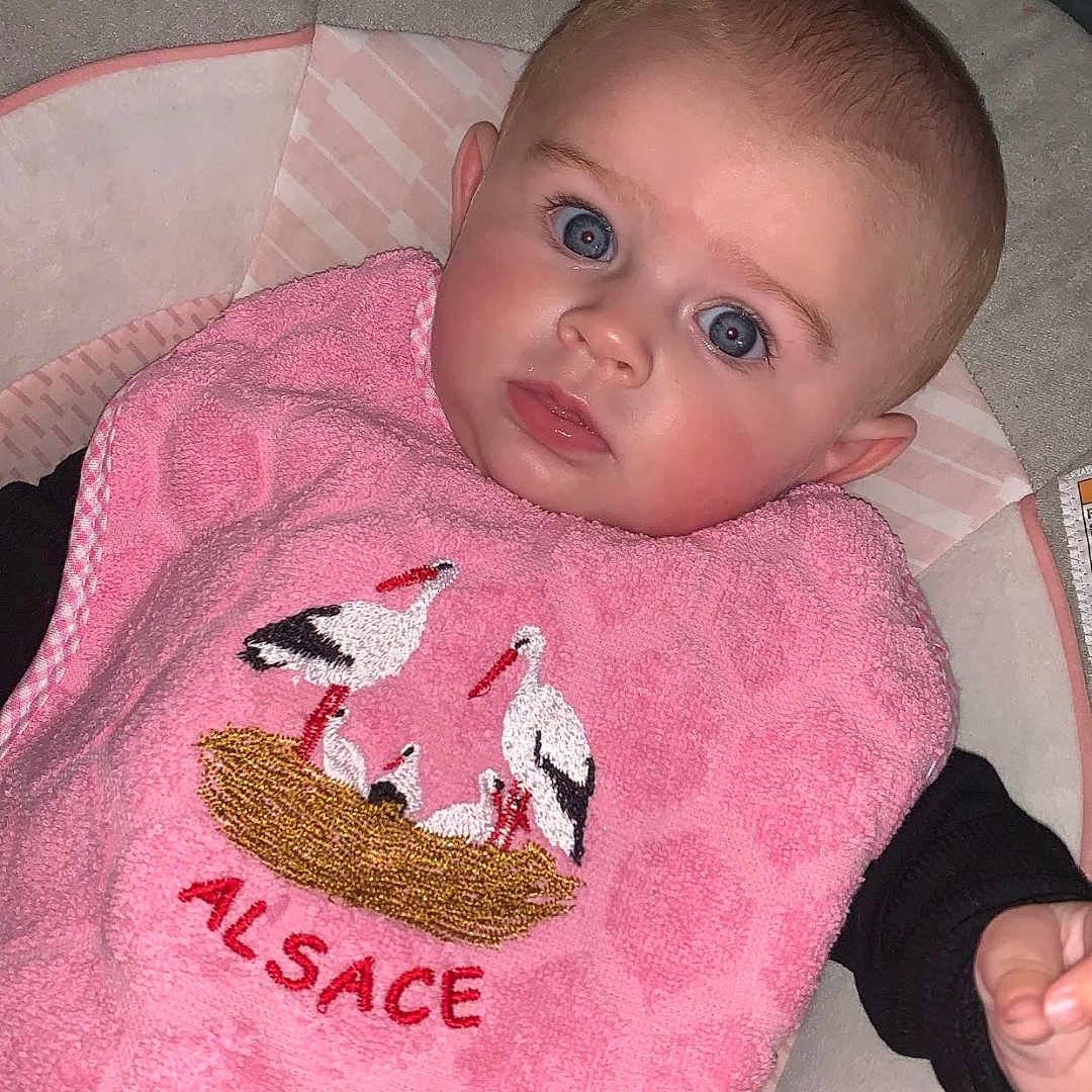 Louise a rejoint le concours — aidez-le/la à gagner de superbes lots ! baby, baby_accessory, bib, blue_eyes, child, close_up, clothing, curious_expression, cushioned_seat, cute, face, hand, indoor, infant, patterned_pants, pink_bib, portrait, seated, soft_lighting, stork_embroidery