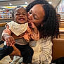 baby, woman, smile, kiss, curly_hair, indoor, restaurant, booth, table, clothing, bib, happy, person, affection, holding, casual_wear, skin, earrings, hair, joy