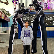 Olivia joined the competition — help win amazing prizes! child, person, skeleton_costume, stilt_walker, hoodie, smile, amusement_park, stage_lighting, performers, costume, outdoor, evening, festival, decorations, band, platform, shoes, concrete_floor, smiling_child, black_clothing