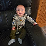 Grayson joined the competition — help win amazing prizes! baby, child, dinosaur_pajamas, sitting, black_leather_chair, happy, smiling, indoor, carpet, wooden_cabinet, pacifier_clip, cute, infant, person, clothing, sock, furniture, portrait, expression, home