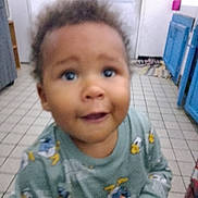 Ryan a rejoint le concours — aidez-le/la à gagner de superbes lots ! toddler, child, indoor, kitchen, sweater, cartoon_print, curly_hair, face, expression, floor_tiles, cabinet, door, home, young_child, person, smile, curious, blurry, casual_clothing, standing