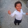 child, smiling, curly_hair, white_shirt, dark_pants, black_shoes, outdoor, pavement, standing, happy, person, casual_clothing, daylight, young_child, motion, arms_raised, sidewalk, playful, cute
