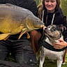 dog, french_bulldog, carp, fish, people, man, woman, outdoors, fishing, angling, large_fish, pet, animal, portrait, cloudy_sky, trees, grass, black_clothing, smiling, text_overlay