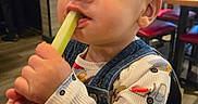 Oliver joined the competition — help win amazing prizes! background, casual, celery, chair, child, eating, expression, face, food, food_item, furniture, hands, high_chair, indoor, overalls, person, restaurant, shirt, table, toddler