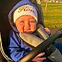 baby, child, stroller, hoodie, knit_hat, blue_clothing, outdoor, sunlight, grass, smile, face, hand, safety_harness, seat, park, daytime, cute, portrait, infant, warm_clothing