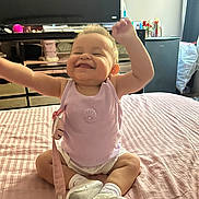 Madisson is registered to the contest to win money with this photo: arms_raised, baby, bed, bedroom, child, clothing, face, happy, home, indoor, joy, onesie, pacifier, pink, plaid, portrait, sitting, smiling, television, toy
