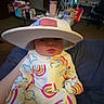 Everleigh is registered to the contest to win money with this photo: baby, child, cowboy_hat, rainbow_outfit, onesie, sitting, portrait, indoors, living_room, couch, television, snacks, table, home, colorful, infant, face, hat, clothing, family_scene