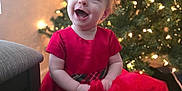 Athena is registered to the contest to win money with this photo: child, toddler, baby, red_dress, christmas_tree, ornament, lights, present, gift, wrapping_paper, smile, joy, holiday, festive, indoor, portrait, sitting, chair, floor, bokeh