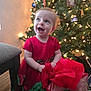 child, toddler, baby, red_dress, christmas_tree, ornament, lights, present, gift, wrapping_paper, smile, joy, holiday, festive, indoor, portrait, sitting, chair, floor, bokeh
