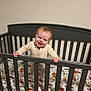baby, infant, child, crib, mattress, bedding, animal_print, smile, happy, standing, onesie, pajamas, nursery, headboard, railing, hands, face, indoor, portrait, wooden_furniture