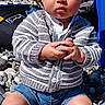toddler, child, outdoor, pebbles, rock, sweater, denim_shorts, black_sneakers, serious_expression, sunny, sky, trees, baby, sitting, holding, casual_clothing, nature, portrait, daytime, young_child