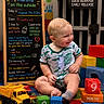 baby, child, toys, blocks, play_mat, signboard, indoor, milestone, smiling, colorful, clothing, floor, person, cute, portrait, sitting, play_jail, text, happy, infant