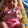 baby, infant, pink_clothing, sitting, person, legs, hands, face, home_interior, furniture, poster, shelf, floral_pattern, yellow_clothing, cute, expression, casual, indoor, relaxed, child
