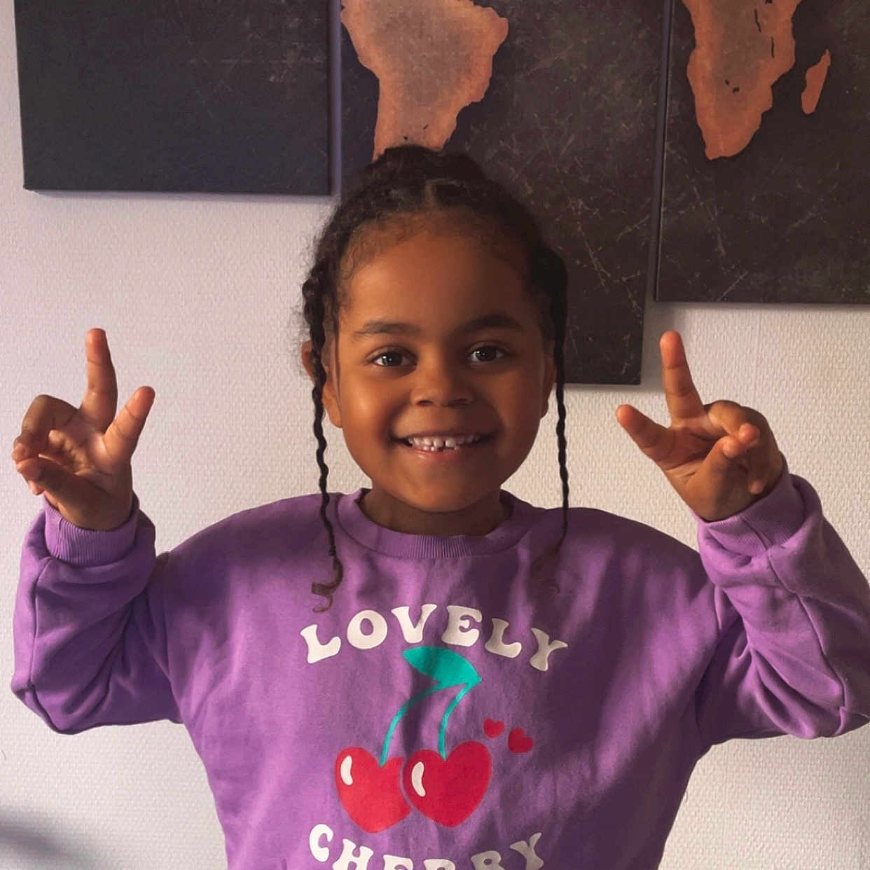 Mya participe au concours pour gagner de l'argent avec cette photo : child, smiling, purple_sweatshirt, cherry_graphic, peace_sign, indoor, world_map, wall_art, happy, person, casual_clothing, braided_hair, portrait, cute, young, playful, fashion, sweatshirt, standing, bright