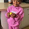 appliance, cabinet, casual_clothing, child, countertop, curly_hair, flooring, happy, indoor, kitchen, map, nike, paper_towel, person, pink_tracksuit, poster, smile, standing, yellow_ball, young