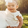 baby, child, smiling, white_bow, headband, outdoor, sunlight, nature, greenery, portrait, happy, infant, holding, adult_hand, white_clothing, cute, earrings, warm_light, person, closeup
