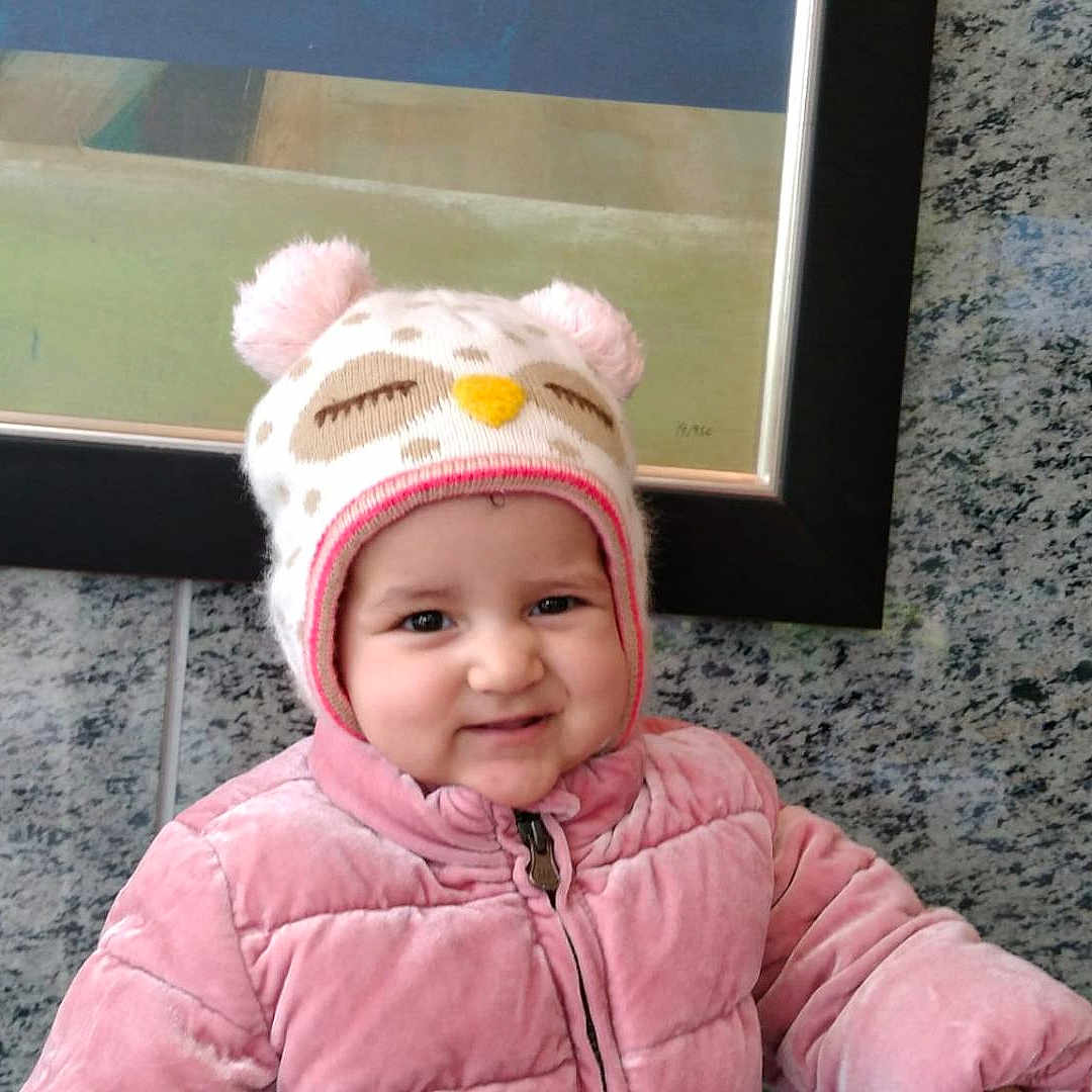 Celia is registered to the contest to win money with this photo: baby, child, infant, portrait, smiling, happy, winter_clothing, pink_jacket, hat, animal_hat, outdoorwear, face, person, indoor, framed_art, wall, granite, sitting, adorable, photograph