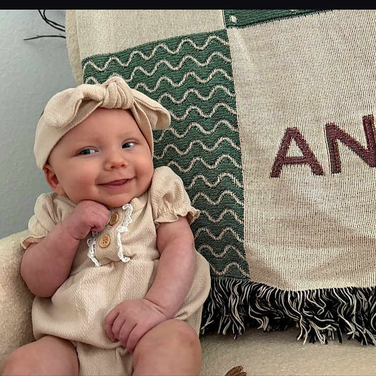 Anya is registered to the contest to win money with this photo: baby, infant, smile, headband, beige_clothing, sitting, chair, blanket, pattern, texture, indoor, cute, portrait, happy, child, person, cozy, fabric, home, comfort