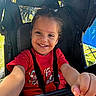 baby, casual_clothing, child, daytime, face, grass, hand, happy, outdoor, person, playful, portrait, reaching, red_shirt, seat, seatbelt, smiling, stroller, sunlight, young