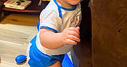 Colter is registered to the contest to win money with this photo: baby, child, smiling, standing, toys, indoors, wooden_floor, couch, happy, cute, playtime, blue_shorts, white_shirt, floor, young_child, exploring, home, colorful, person, infant