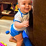 baby, child, smiling, standing, toys, indoors, wooden_floor, couch, happy, cute, playtime, blue_shorts, white_shirt, floor, young_child, exploring, home, colorful, person, infant