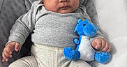 Ronn is registered to the contest to win money with this photo: baby, baby_toys, blue_dragon, child, comfort, cream_pants, cushioned_seat, cute, face, gray_clothing, hand, indoors, infant, infant_clothing, person, plush_toy, portrait, seated, soft_fabric, young_child