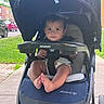 baby, stroller, child, outdoor, sidewalk, grass, car, curious, seatbelt, foot, hand, canopy, daylight, person, urban, parked_car, smile, infant, transportation, casual