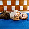 baby, child, smiling, bed, blue_bedspread, brown_pants, shirtless, wooden_headboard, indoor, happy, infant, lying_down, cute, portrait, playful, home, person, young_child, sleepwear, relaxed
