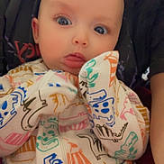 Nayeli joined the competition — help win amazing prizes! baby, infant, child, face, big_eyes, tongue_out, onesie, dinosaur_print, patterned_clothing, hands_near_face, cheeks, closeup, portrait, sitting, parent_arm, blanket, cozy, indoor, cute, wide_eyes