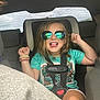 child, car_seat, sunglasses, car_interior, seatbelt, happy, playful, smiling, tongue_out, casual_clothing, bracelets, hair, seat, window, reflection, person, kid, safety, fun, transportation