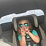 child, car_seat, sunglasses, peace_sign, car_interior, seatbelt, wristbands, turquoise_shirt, smiling, happy, young_child, reflection, casual_clothing, sitting, vehicle, backseat, playful, blond_hair, daylight, portrait