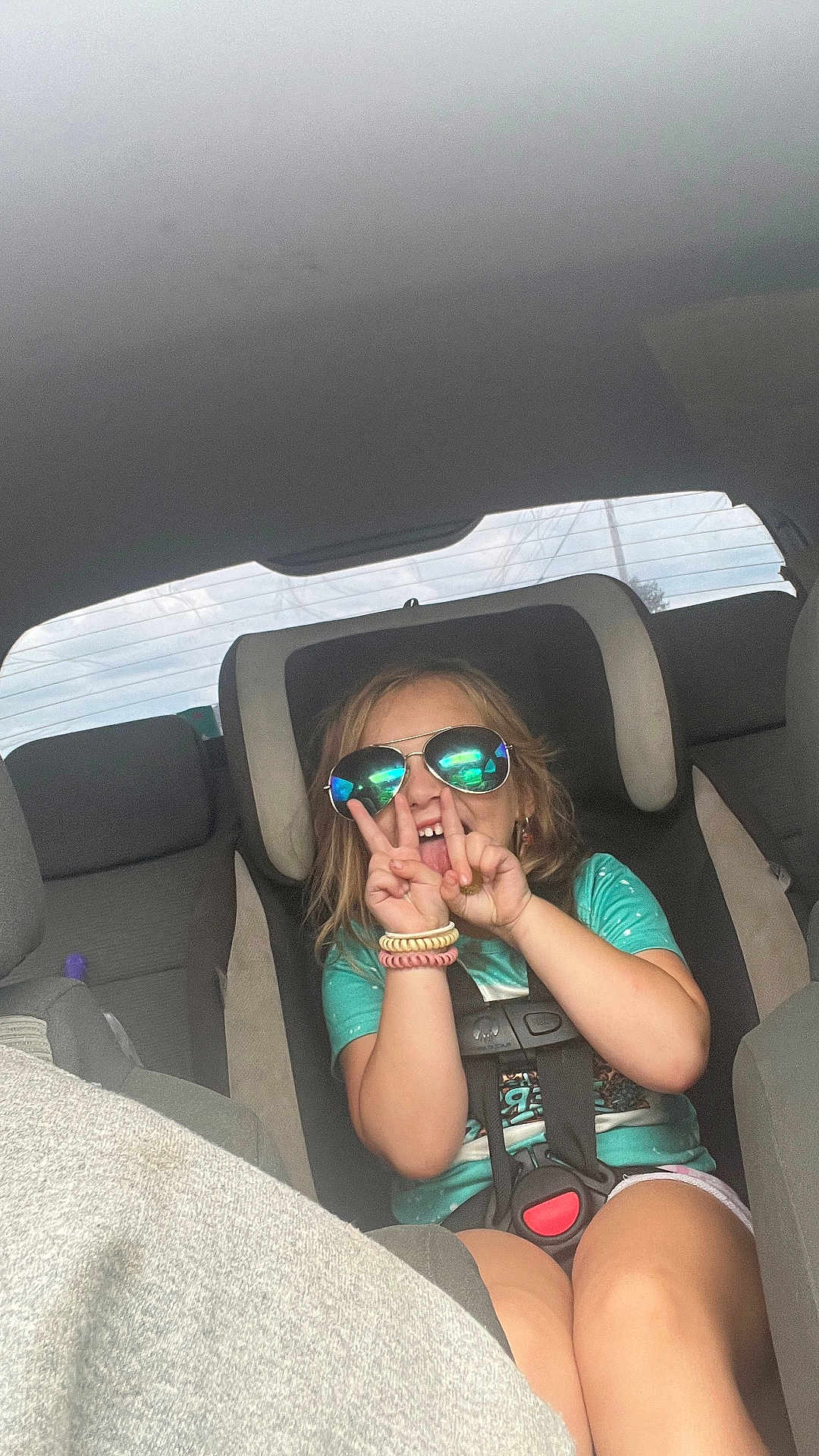 Ophelia-Rayne joined the competition — help win amazing prizes! child, car_seat, sunglasses, peace_sign, car_interior, seatbelt, wristbands, turquoise_shirt, smiling, happy, young_child, reflection, casual_clothing, sitting, vehicle, backseat, playful, blond_hair, daylight, portrait