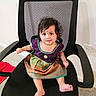baby, child, office_chair, mesh_chair, colorful_dress, traditional_clothing, barefoot, indoor, carpet, wall, power_outlet, cute, sitting, looking_up, black_chair, curly_hair, infant, young_child, adorable, portrait
