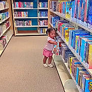 Hosmi joined the competition — help win amazing prizes! toddler, child, library, bookshelf, books, carpet, indoor, curious, reading, young_child, pink_shorts, white_shoes, aisle, education, learning, literature, small_person, floor, shelves, colorful