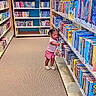 toddler, child, library, bookshelf, books, carpet, indoor, curious, reading, young_child, pink_shorts, white_shoes, aisle, education, learning, literature, small_person, floor, shelves, colorful