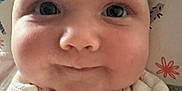 Iris is registered to the contest to win money with this photo: baby, face, close_up, child, infant, cheeks, eyes, knitted_clothing, white_clothing, pillow, patterned_pillow, floral_pattern, cute, young_child, skin, portrait, indoors, smile, head, expression